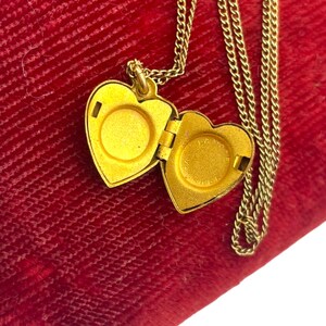 May include: A gold-colored heart-shaped locket necklace with an open compartment. The locket hangs on a delicate chain, resting on a red velvet surface. The locket is open, revealing the interior. The necklace is a piece of jewelry.