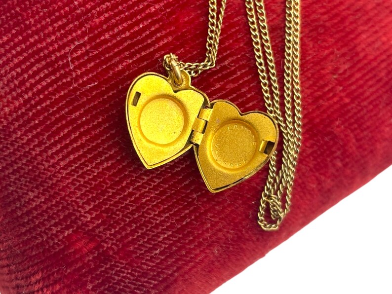 May include: A gold-colored heart-shaped locket necklace with an open compartment. The locket hangs on a delicate chain, resting on a red velvet surface. The locket is open, revealing the interior. The necklace is a piece of jewelry.