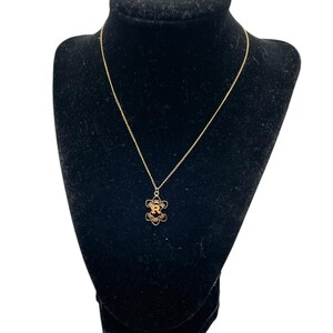 May include: A gold-colored necklace with a floral pendant featuring the letter "R" in the center. The necklace is displayed on a black velvet bust, showcasing the delicate chain and pendant design. The background is white.