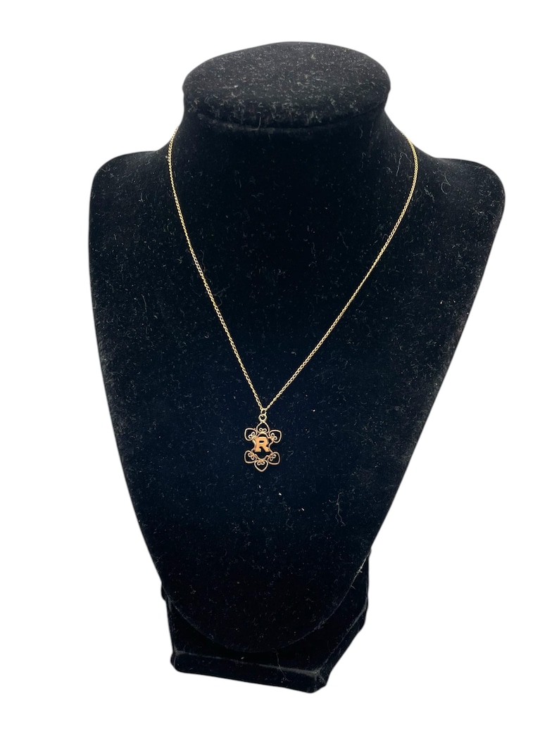 May include: A gold-colored necklace with a floral pendant featuring the letter "R" in the center. The necklace is displayed on a black velvet bust, showcasing the delicate chain and pendant design. The background is white.