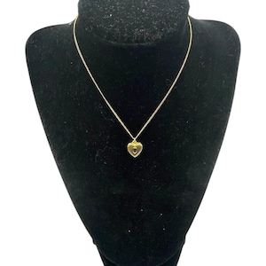 May include: A gold-colored necklace with a heart-shaped pendant, displayed on a black velvet bust. The pendant has a small, circular design in the center. The chain is delicate and thin.