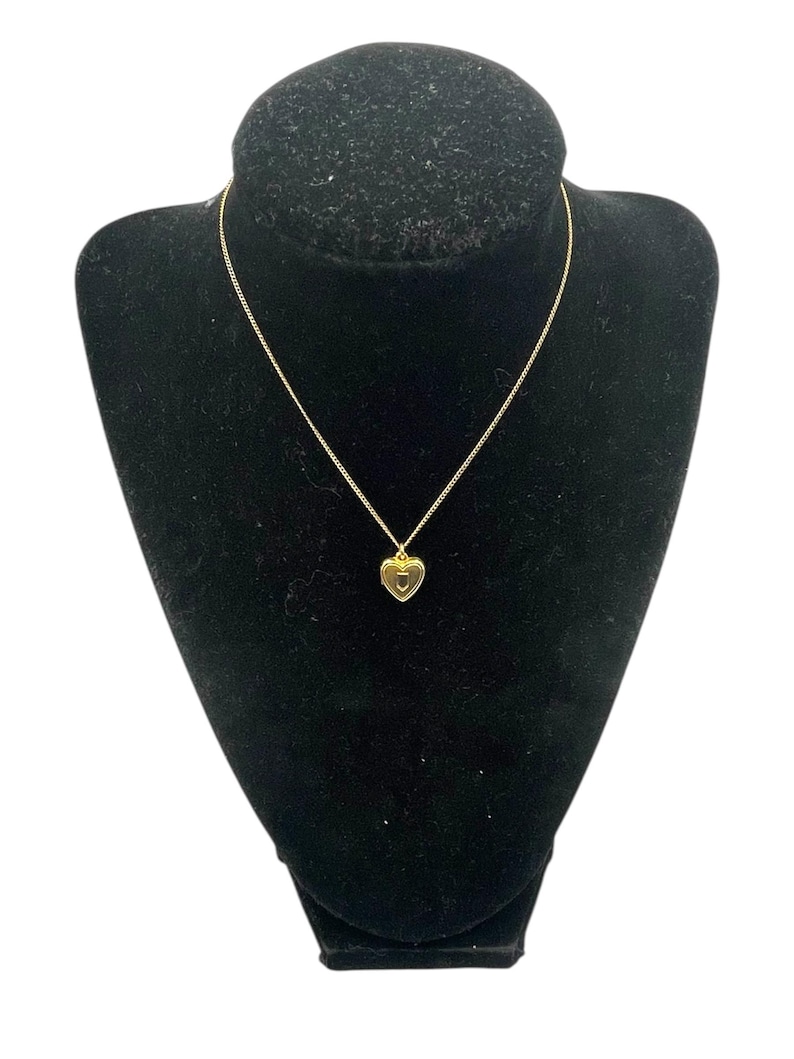 May include: A gold-colored necklace with a heart-shaped pendant, displayed on a black velvet bust. The pendant has a small, circular design in the center. The chain is delicate and thin.