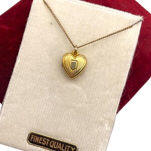 May include: A gold-colored heart-shaped pendant necklace. The pendant has a shield design in the center and is on a delicate chain. The necklace is presented on a white velvet card with a "FINEST QUALITY" label, set against a red velvet background.