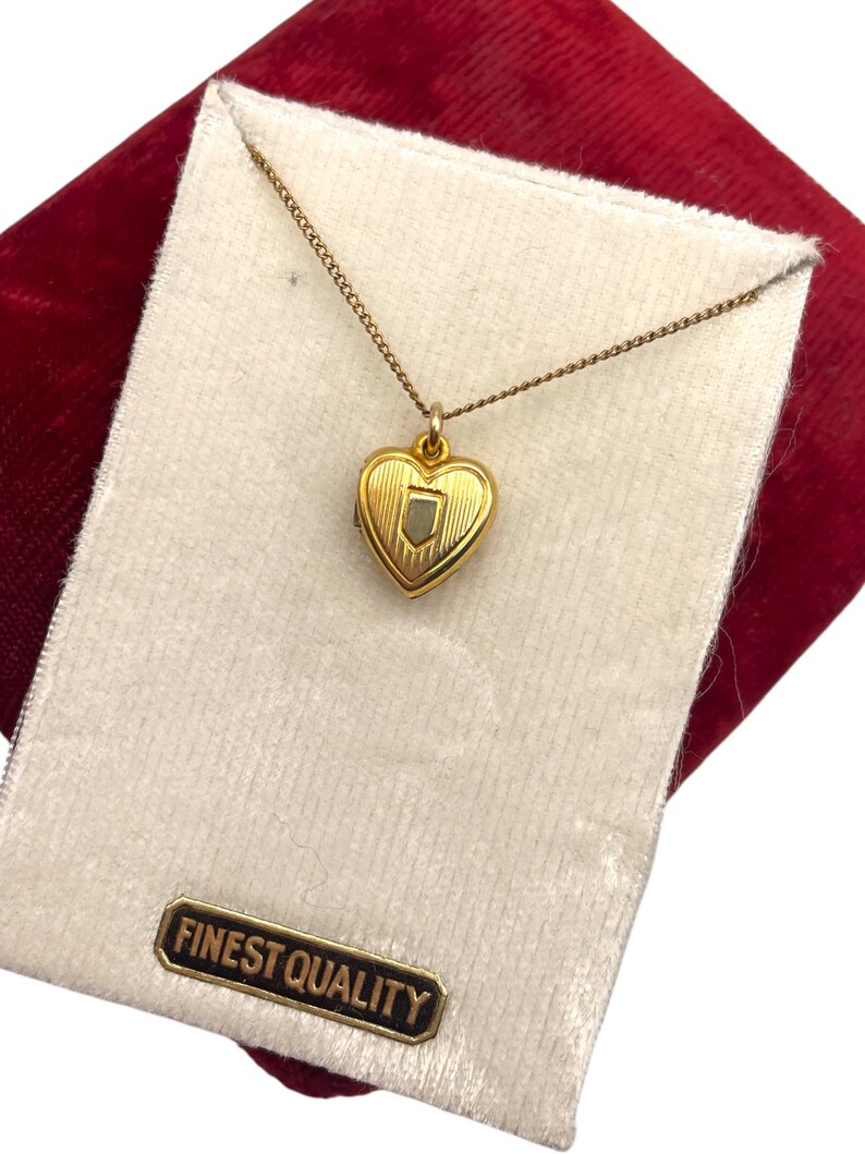 May include: A gold-colored heart-shaped pendant necklace. The pendant has a shield design in the center and is on a delicate chain. The necklace is presented on a white velvet card with a "FINEST QUALITY" label, set against a red velvet background.