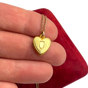 May include: A gold-colored heart-shaped locket pendant on a delicate chain. The locket features a shield design on the front and is held against a red background. The jewelry is likely vintage or antique.