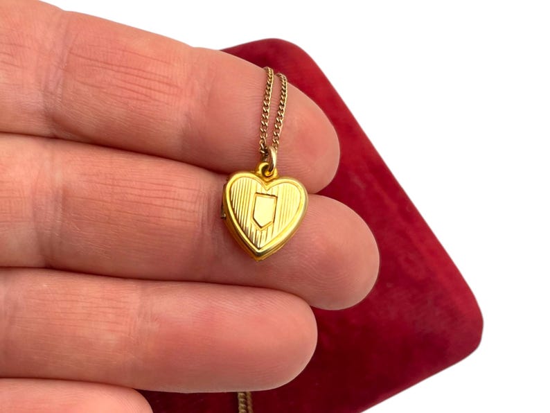 May include: A gold-colored heart-shaped locket pendant on a delicate chain. The locket features a shield design on the front and is held against a red background. The jewelry is likely vintage or antique.