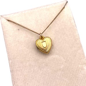 May include: A gold-colored heart-shaped locket necklace with a small shield design. The locket hangs from a delicate chain and is displayed on a light pink velvet-like material. The locket has a textured surface and a small clasp.