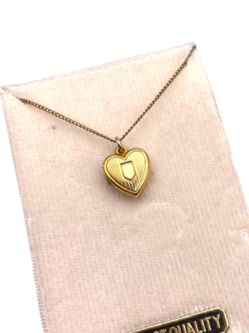 May include: A gold-colored heart-shaped locket necklace with a small shield design. The locket hangs from a delicate chain and is displayed on a light pink velvet-like material. The locket has a textured surface and a small clasp.