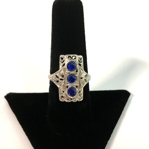 May include: A silver ring with three bright blue gemstones. The ring has an ornate filigree design around the stones. It is displayed on a black stand. The ring's intricate details are highlighted against the black background.