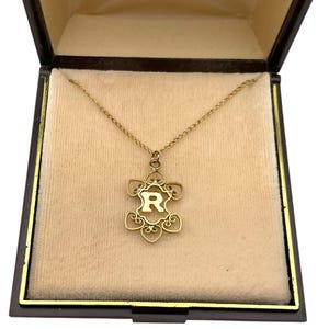 May include: A gold-colored necklace with a pendant shaped like a stylized flower, featuring the letter "R" in the center. The necklace is displayed inside a black jewelry box with a beige interior, creating a luxurious presentation.