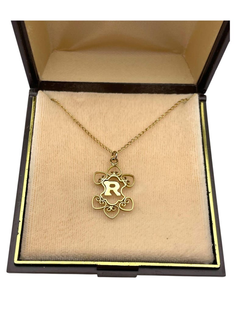 May include: A gold-colored necklace with a pendant shaped like a stylized flower, featuring the letter "R" in the center. The necklace is displayed inside a black jewelry box with a beige interior, creating a luxurious presentation.
