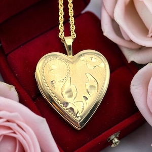 May include: A gold heart-shaped locket pendant with an engraved floral design, displayed in a red velvet jewelry box. The pendant hangs from a delicate gold chain. Pink roses are in the background.
