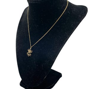 May include: A gold-colored necklace with a floral pendant is displayed on a black velvet bust. The pendant features a delicate, openwork design with small, round embellishments. The chain is fine and elegant, complementing the pendant's intricate details.