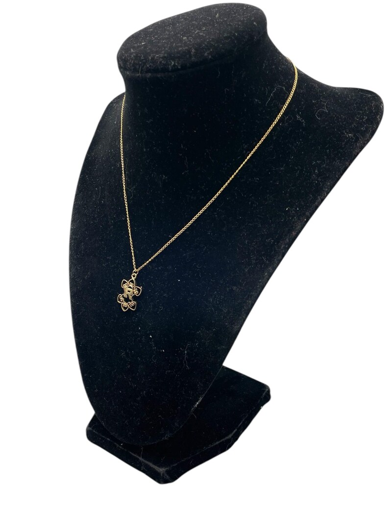 May include: A gold-colored necklace with a floral pendant is displayed on a black velvet bust. The pendant features a delicate, openwork design with small, round embellishments. The chain is fine and elegant, complementing the pendant's intricate details.