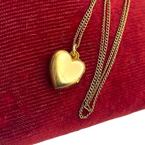 May include: A gold heart-shaped locket necklace on a red velvet surface. The locket is suspended from a delicate gold chain. The heart locket is closed and has a smooth, reflective surface.