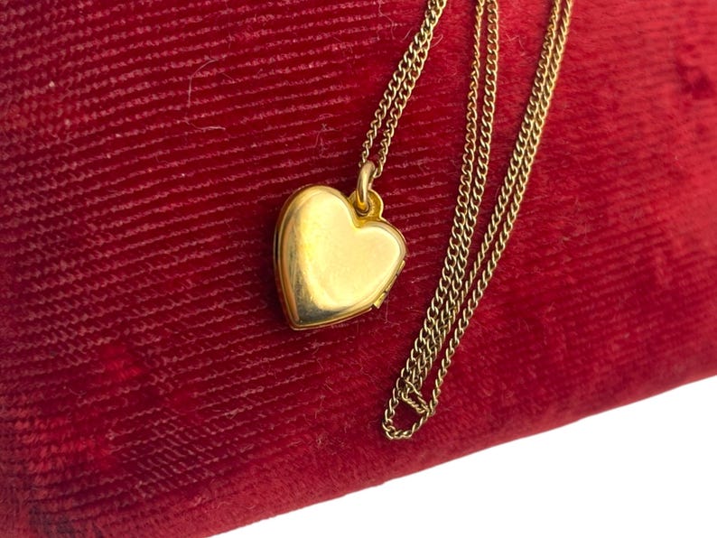 May include: A gold heart-shaped locket necklace on a red velvet surface. The locket is suspended from a delicate gold chain. The heart locket is closed and has a smooth, reflective surface.