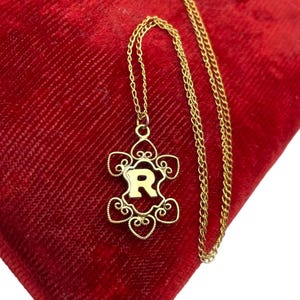 May include: A gold-colored necklace with a pendant featuring the letter "R" in a decorative, openwork design. The pendant is suspended from a delicate chain, resting on a red velvet surface. The necklace is a jewelry item.