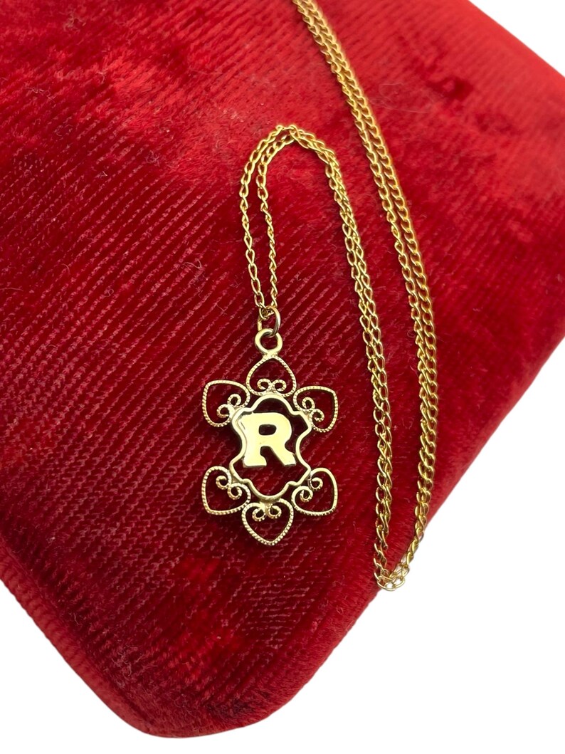 May include: A gold-colored necklace with a pendant featuring the letter "R" in a decorative, openwork design. The pendant is suspended from a delicate chain, resting on a red velvet surface. The necklace is a jewelry item.