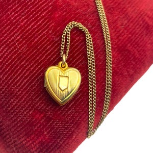 May include: A gold-colored heart-shaped locket pendant with a shield design, suspended from a delicate chain. The locket rests on a red velvet surface, creating a striking contrast. The chain is approximately 18 inches long.