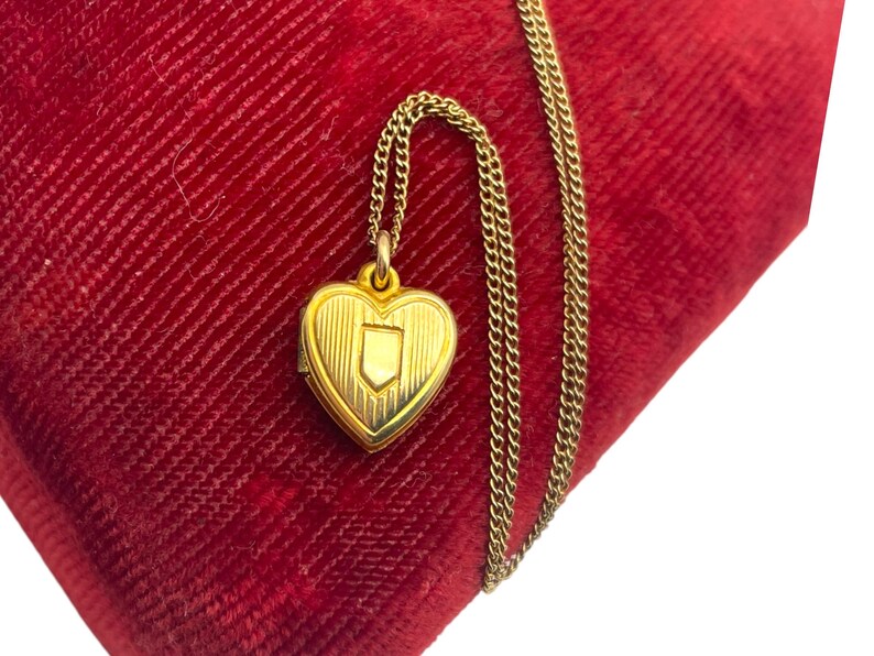 May include: A gold-colored heart-shaped locket pendant with a shield design, suspended from a delicate chain. The locket rests on a red velvet surface, creating a striking contrast. The chain is approximately 18 inches long.
