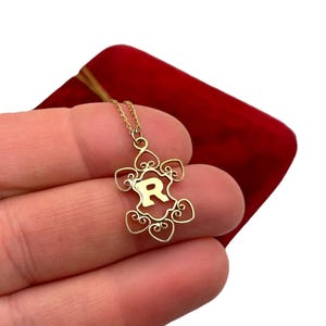 May include: A gold-colored pendant necklace featuring the letter "R" within a decorative frame. The pendant hangs from a delicate chain. The necklace is displayed against a red velvet background.