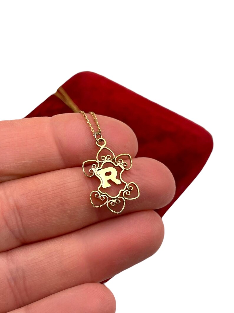 May include: A gold-colored pendant necklace featuring the letter "R" within a decorative frame. The pendant hangs from a delicate chain. The necklace is displayed against a red velvet background.