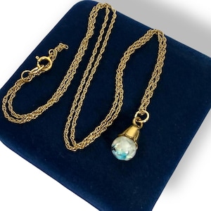 May include: A gold-toned necklace with a delicate chain and a pendant. The pendant is a small, round glass orb with blue and white accents. The necklace is displayed on a dark blue velvet surface.