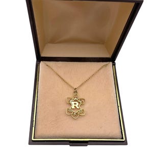 May include: A gold-colored necklace with a floral pendant containing the letter "R" inside a dark brown jewelry box with a beige interior. The necklace is displayed on a beige velvet-like surface.