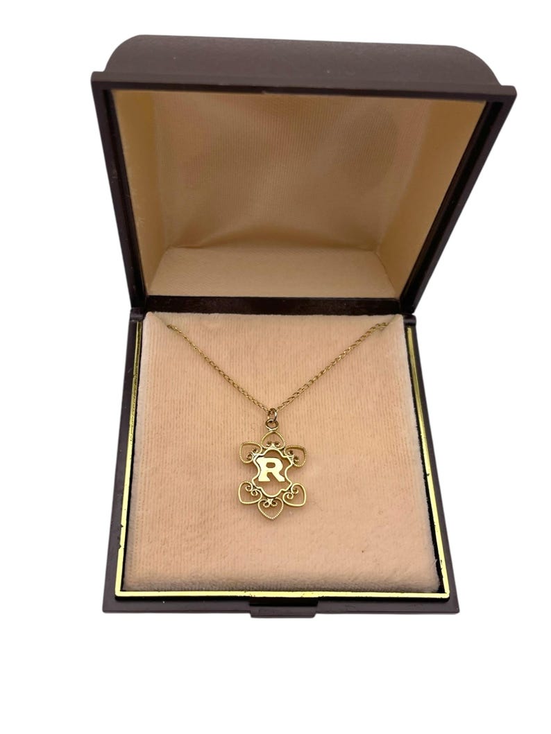 May include: A gold-colored necklace with a floral pendant containing the letter "R" inside a dark brown jewelry box with a beige interior. The necklace is displayed on a beige velvet-like surface.