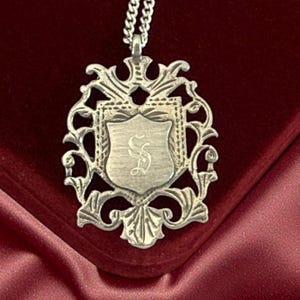 Puede incluir: Collar con colgante plateado con un diseño ornamentado en forma de escudo. El colgante presenta un escudo central con una inicial grabada, rodeado de volutas decorativas y una cadena. El collar se muestra sobre una superficie de terciopelo burdeos.