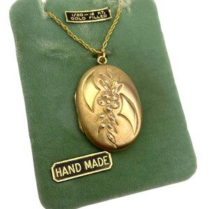 May include: Gold-filled oval locket necklace with floral design, displayed on a green velvet card. The locket is suspended from a delicate chain. The card has a label that says "HAND MADE".