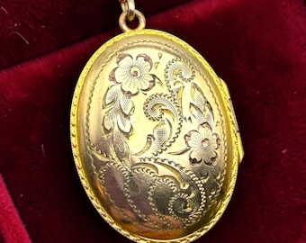 Puede incluir: Un colgante de relicario ovalado de oro suspendido de una cadena de cuerda de oro a juego. El relicario presenta un intrincado patrón floral grabado con hojas y flores en espiral en la parte frontal, enmarcado por un borde decorativo.