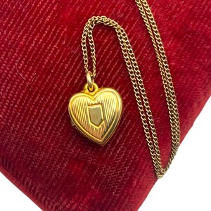 May include: A gold-colored heart-shaped locket pendant with a shield design, suspended from a delicate chain. The locket rests on a plush, red velvet surface, creating a striking contrast. The chain and locket appear to be made of a gold-toned metal.