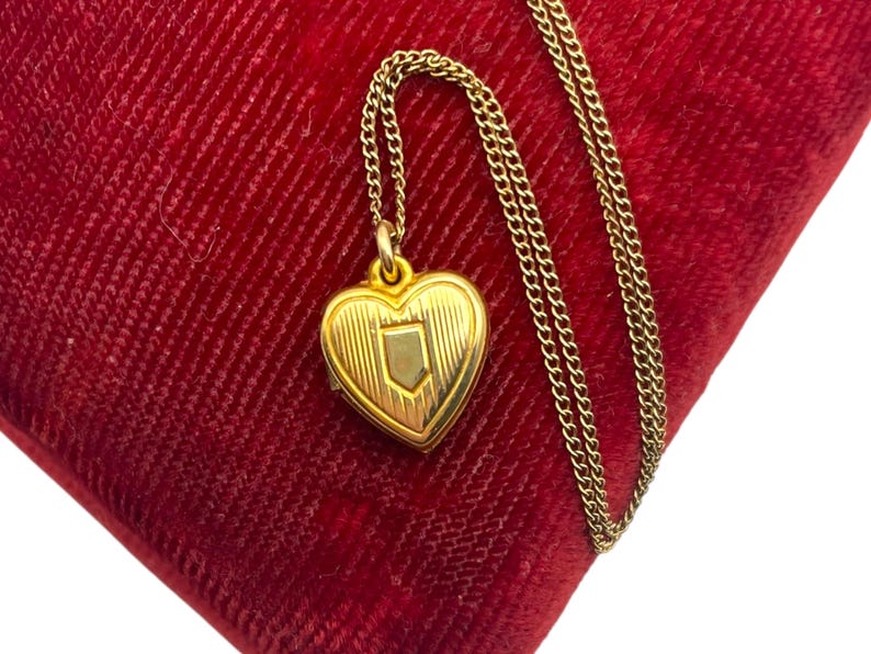 May include: A gold-colored heart-shaped locket pendant with a shield design, suspended from a delicate chain. The locket rests on a plush, red velvet surface, creating a striking contrast. The chain and locket appear to be made of a gold-toned metal.