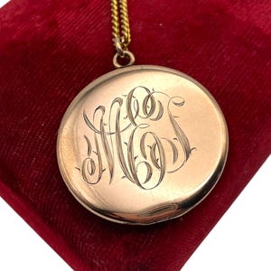 May include: A gold-colored round locket pendant with an engraved monogram, suspended from a delicate gold chain. The locket rests on a deep red velvet surface, creating a striking contrast. The monogram is in an elegant script.