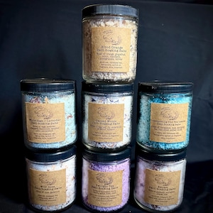 May include: A collection of eight glass jars filled with bath salts. Each jar has a brown paper label with a different scent and name, such as "Blood Orange", "White Sage + Lavender", "Sacred Woods", "Interpreting the Tea Leaves", "Wild Rose", "Twilight Tea", and "Karma".