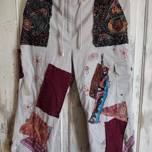 May include: Beige capri pants with decorative patches and embroidered floral and mushroom designs. The patches feature paisley and patterned fabrics in shades of burgundy, brown, and blue. The pants have a drawstring waist and a cargo pocket.