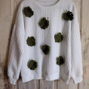 May include: White sweatshirt with a textured cable knit design and olive green crocheted leaf appliqués. The crew neck sweatshirt has long sleeves and a ribbed hem and cuffs. The leaves are evenly spaced across the front of the sweater.