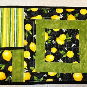 May include: A black quilted placemat with a green border and a lemon print. The center of the placemat has a smaller green border with a lemon print.