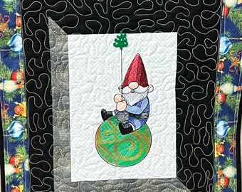 May include: A Christmas-themed quilt featuring a gnome sitting on a green ornament. The gnome wears a red hat and blue jacket. The quilt has a black and white patterned border and a blue border with Christmas ornaments.