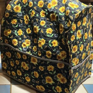 May include: A quilted appliance cover featuring a vibrant sunflower pattern on a black background. The cover has a rounded top and two pockets, with a gray trim. The sunflowers are yellow with brown centers and green leaves.