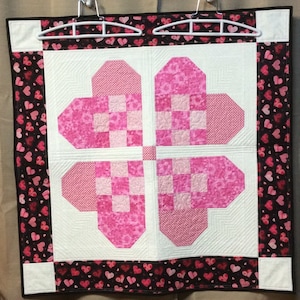 May include: A pink and white quilt with a four-leaf clover design made of hearts. The quilt is surrounded by a black border with a pattern of small pink hearts.