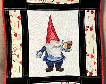 May include: A quilted wall hanging featuring a central gnome wearing a red hat and blue apron, holding a loaf of bread. The border includes black fabric and cream-colored panels printed with red hearts, kitchen utensils, and the text Kiss the Cook.