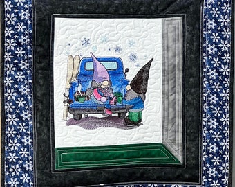 May include: A quilted wall hanging with a winter scene. The central image depicts two gnomes near a blue truck, with skis and snowflakes. The border features a dark blue fabric with white snowflakes.