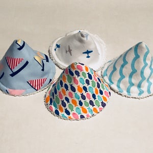 Puede incluir: Cuatro baberos de bebé en forma de cono con diferentes patrones. Los patrones incluyen veleros, aviones, peces y olas. Los baberos son blancos con un borde azul.