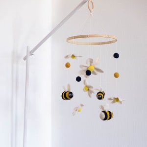 May include: A white wooden baby crib mobile with a yellow and black bee, white felt flowers, and yellow and navy blue pom-poms.