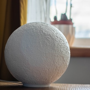 May include: A white moon-shaped lamp with a textured surface sits on a wooden table.