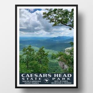 May include: A framed vertical poster featuring a scenic landscape view of Caesars Head State Park in South Carolina. The image displays lush green trees, rocky cliffs, and distant blue mountains under a cloudy sky, with text at the bottom.