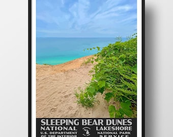 May include: A framed poster featuring a landscape of Sleeping Bear Dunes National Lakeshore with sandy dunes, green vegetation, and a blue lake under a cloudy sky. The text reads SLEEPING BEAR DUNES NATIONAL LAKESHORE U.S. DEPARTMENT OF THE INTERIOR.