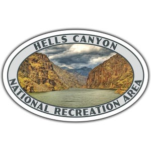 May include: Oval sticker featuring a scenic view of Hells Canyon National Recreation Area. The image shows a river flowing between canyon walls under a cloudy sky. The text "HELLS CANYON NATIONAL RECREATION AREA" is printed around the oval.